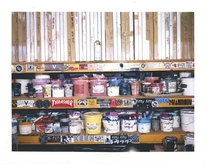 Polaroid of the studio shelves: stacks of screens above rows of ink tubs, every shelf edge covered in stickers