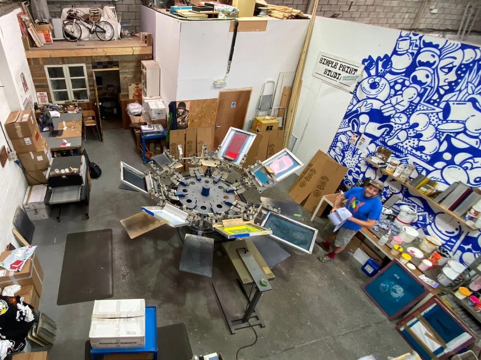 Looking down on the studio: an eight-arm carousel press loaded with screens, a cobalt blue cartoon mural and the printer grinning up at the camera